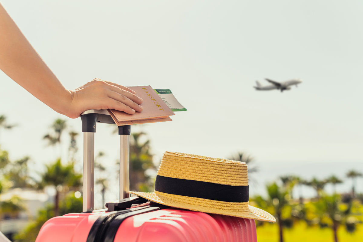 a hand holding a passport and a hat next to a suitcase